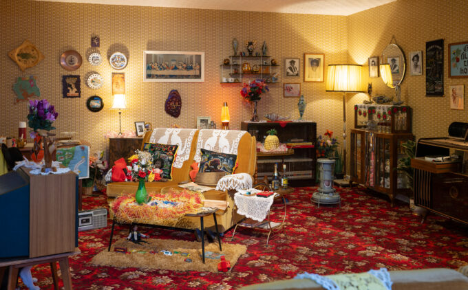 A cluttered 1970s-style living space with yellow wallpaper, red patterned carpet, and furniture and decorations.