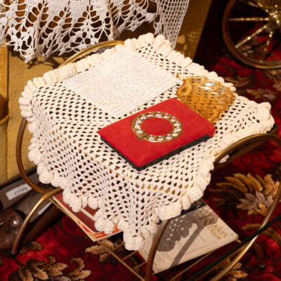 A letter and red item sit on a white lace doily, on a brass table, on a red, floral carpet.