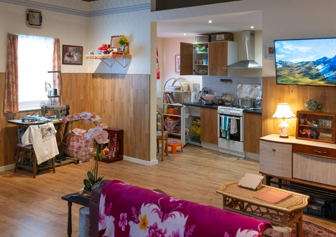 A space showing living room with floral throw over a sofa and connecting kitchen, featuring wood paneling, sewing machine, and wall-mounted TV.