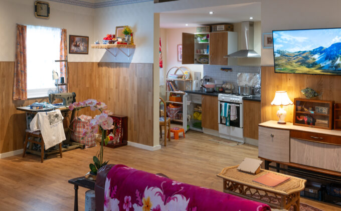 A space showing living room with floral throw over a sofa and connecting kitchen, featuring wood paneling, sewing machine, and wall-mounted TV.