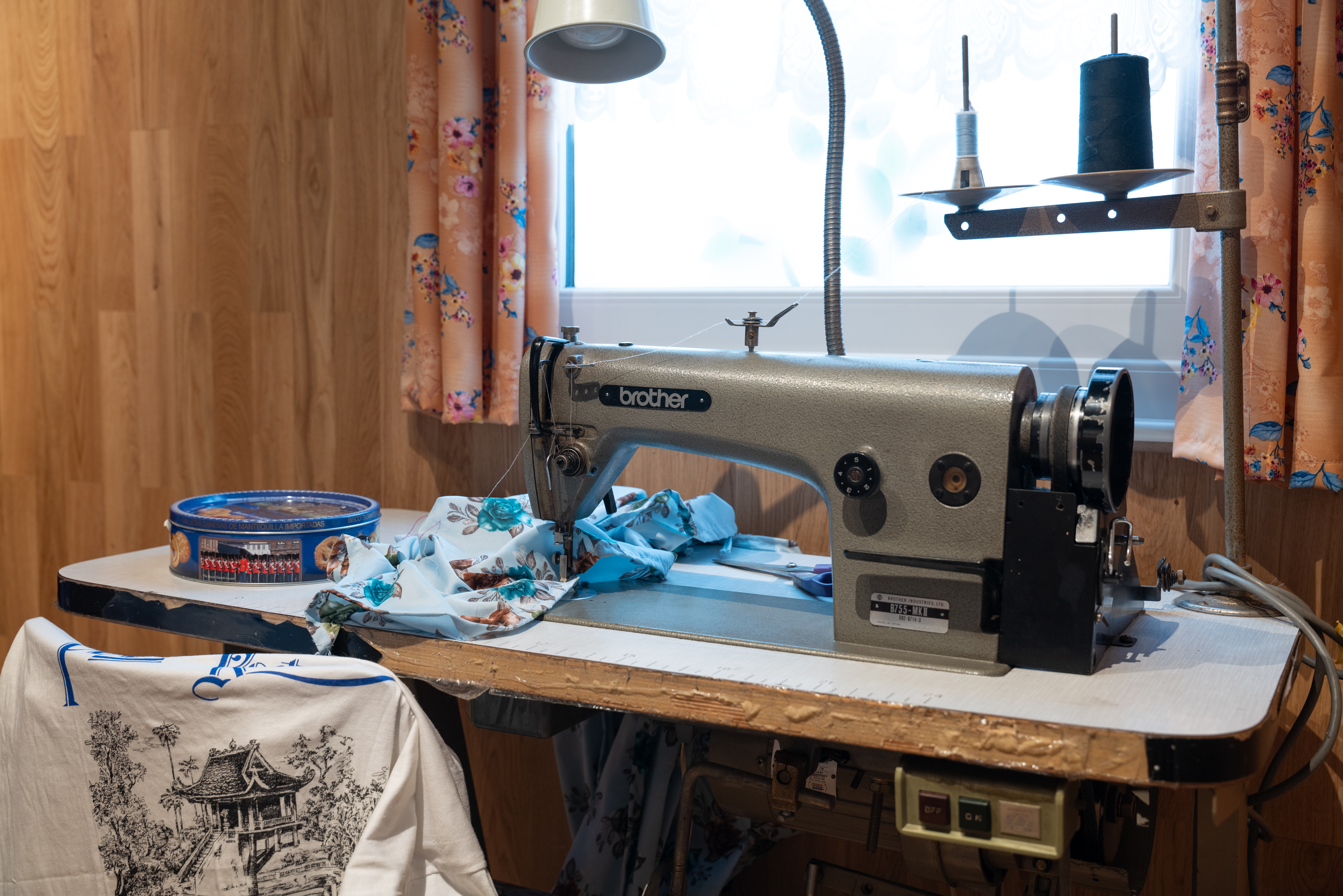 A Brother sewing machine set up on a white table in front of a window, with light blue fabric underneath the needle. The workspace includes a tin of sewing supplies.