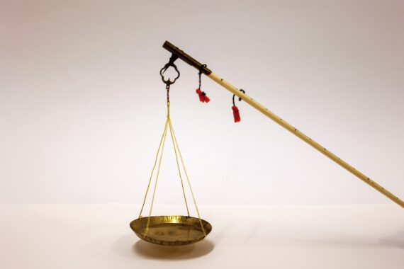 Brass weighing scale with a shallow dish suspended by yellow strings from a long wooden pole with red tassels.