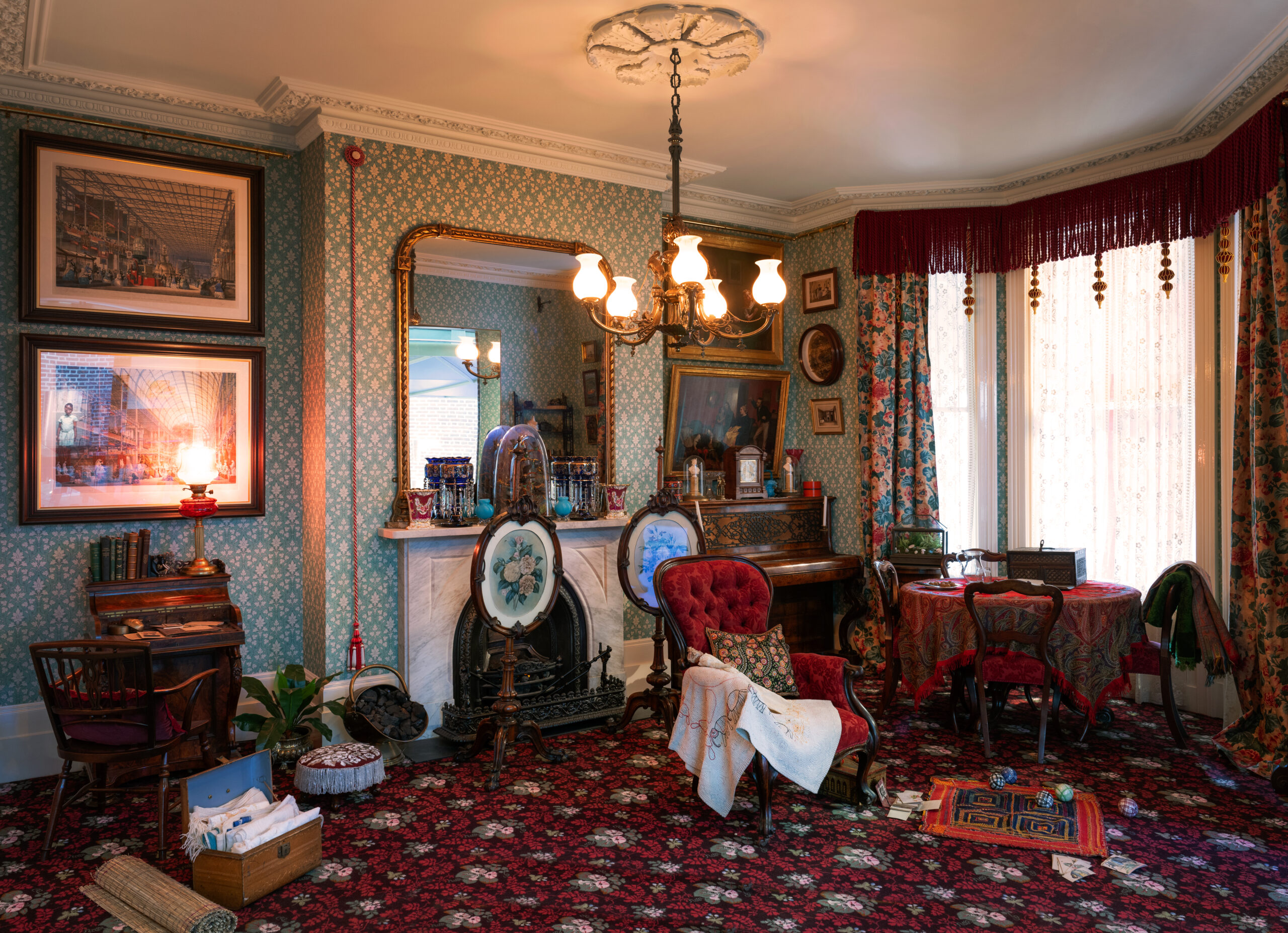 A richly furnished room with mint green patterned wallpaper and red carpet, featuring a red upholstered armchair draped with embroidery beside a marble fireplace topped by twin ceramic vases, framed artwork, a carved wooden piano, a table covered in a red, detailed cloth, and in the left bottom corner is an ayah's trunk of Indian textiles.