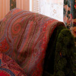 Red paisley-patterned pashmina draped over green velvet chair against floral curtains.