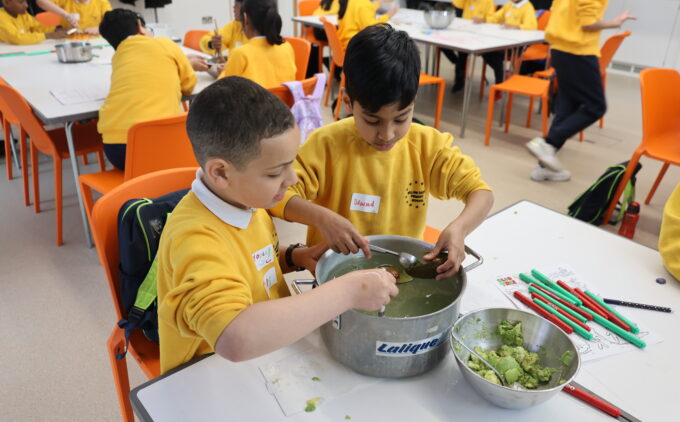 Two primary school students in yellow uniforms stir plant material in a metal tub with spoons, beside a bowl of mashed avocado and colored pens on the table, while classmates work at tables in the background.