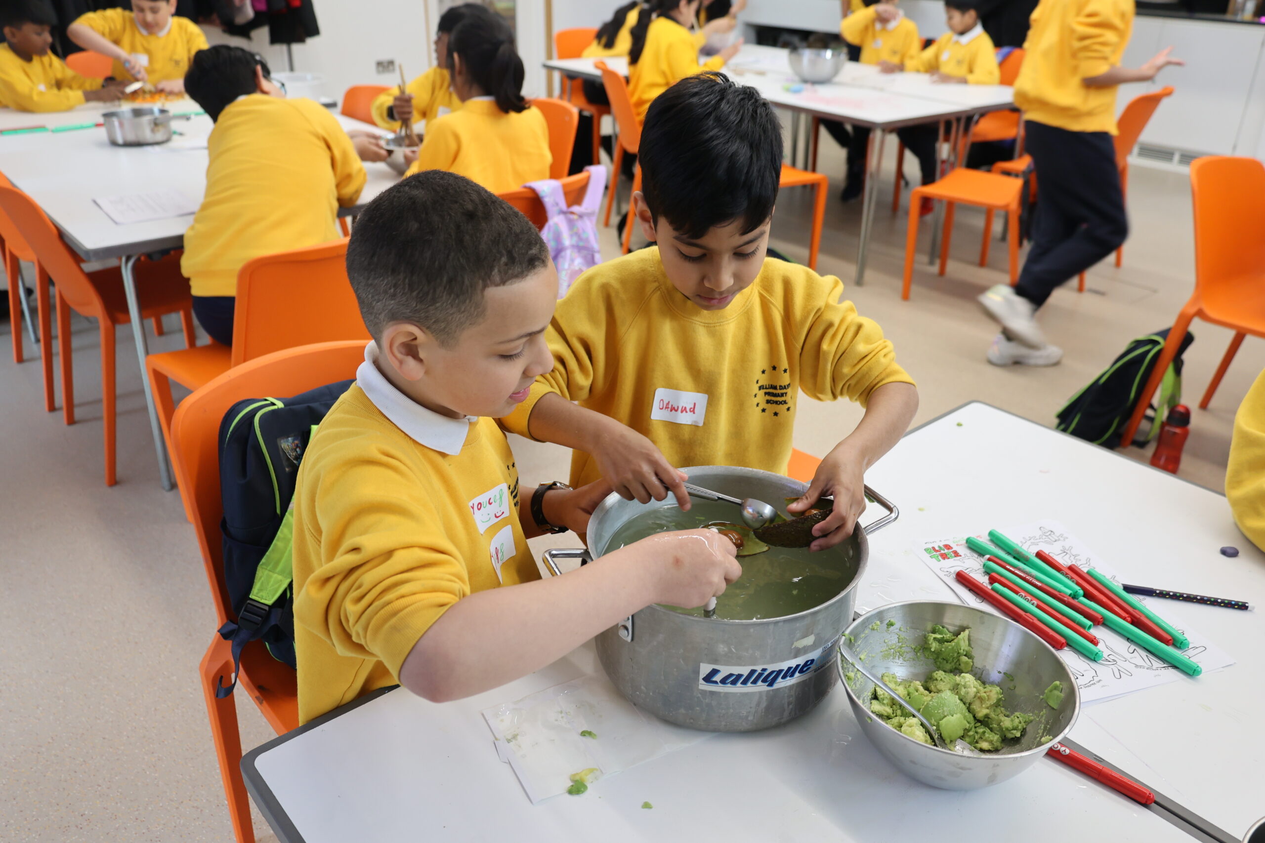 Two primary school students in yellow uniforms stir plant material in a metal tub with spoons, beside a bowl of mashed avocado and colored pens on the table, while classmates work at tables in the background.
