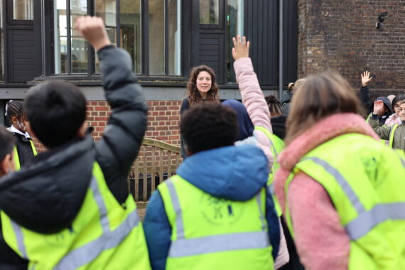 A teacher standing speaking to children, their hands up in the air to ask her a question