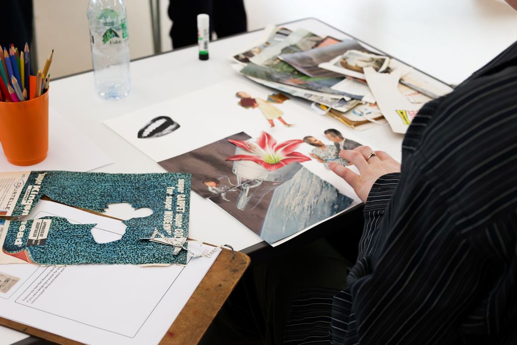 A person's hands arrange cut-out images—flowers, people, and abstract shapes—on a collage sheet at workshop table strewn with pencils, glue sticks, and paper scraps.