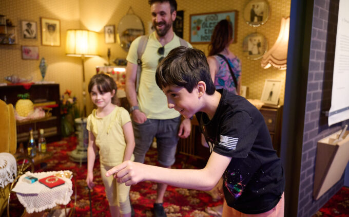 A family in A Terraced House in 1978 installation at the Museum; a smiling child leans forward to point at something outside the frame while the other child and one of the adults watch with smile.