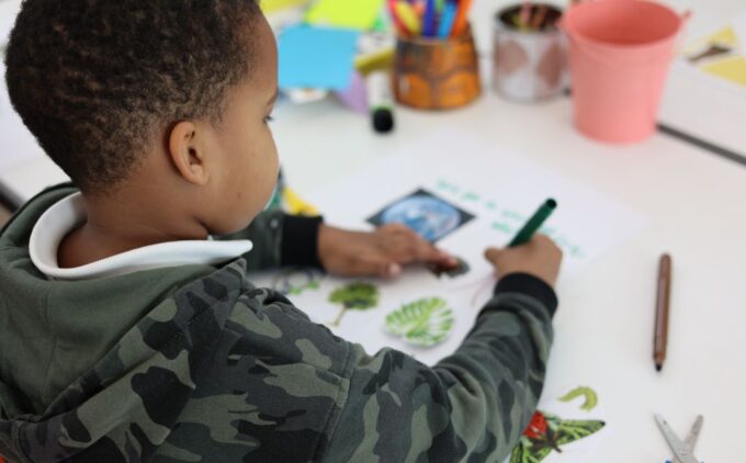 A primary school age child in a camouflage hoodie uses a green marker to write and draw on a paper decorated with cut-out images of plants, surrounded by scissors, colored pens, and craft supplies.
