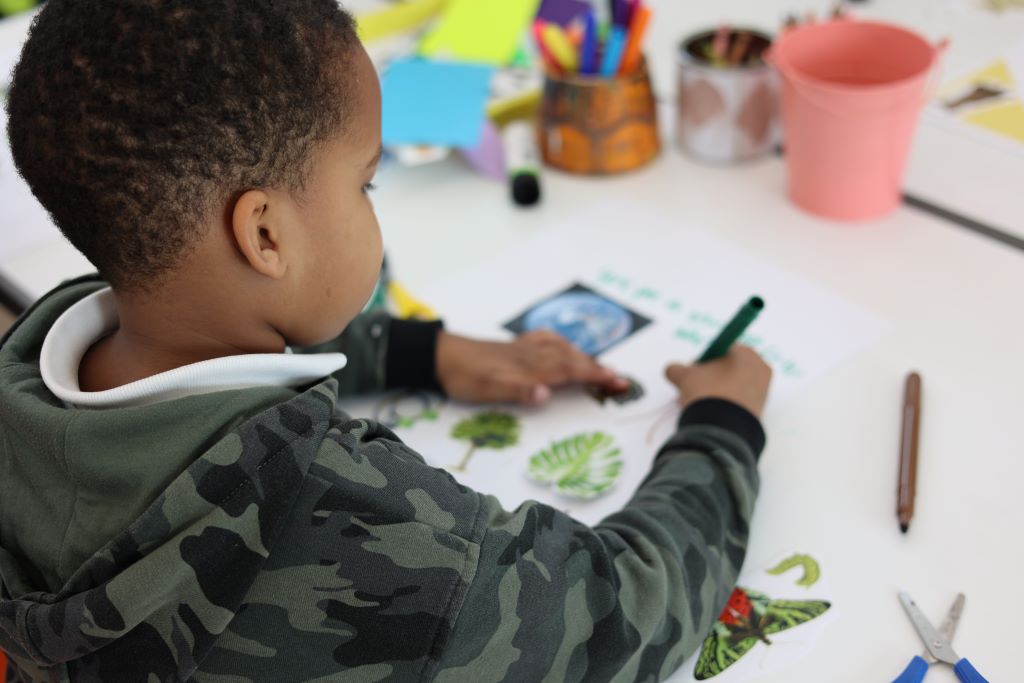 A primary school age child in a camouflage hoodie uses a green marker to write and draw on a paper decorated with cut-out images of plants, surrounded by scissors, colored pens, and craft supplies.