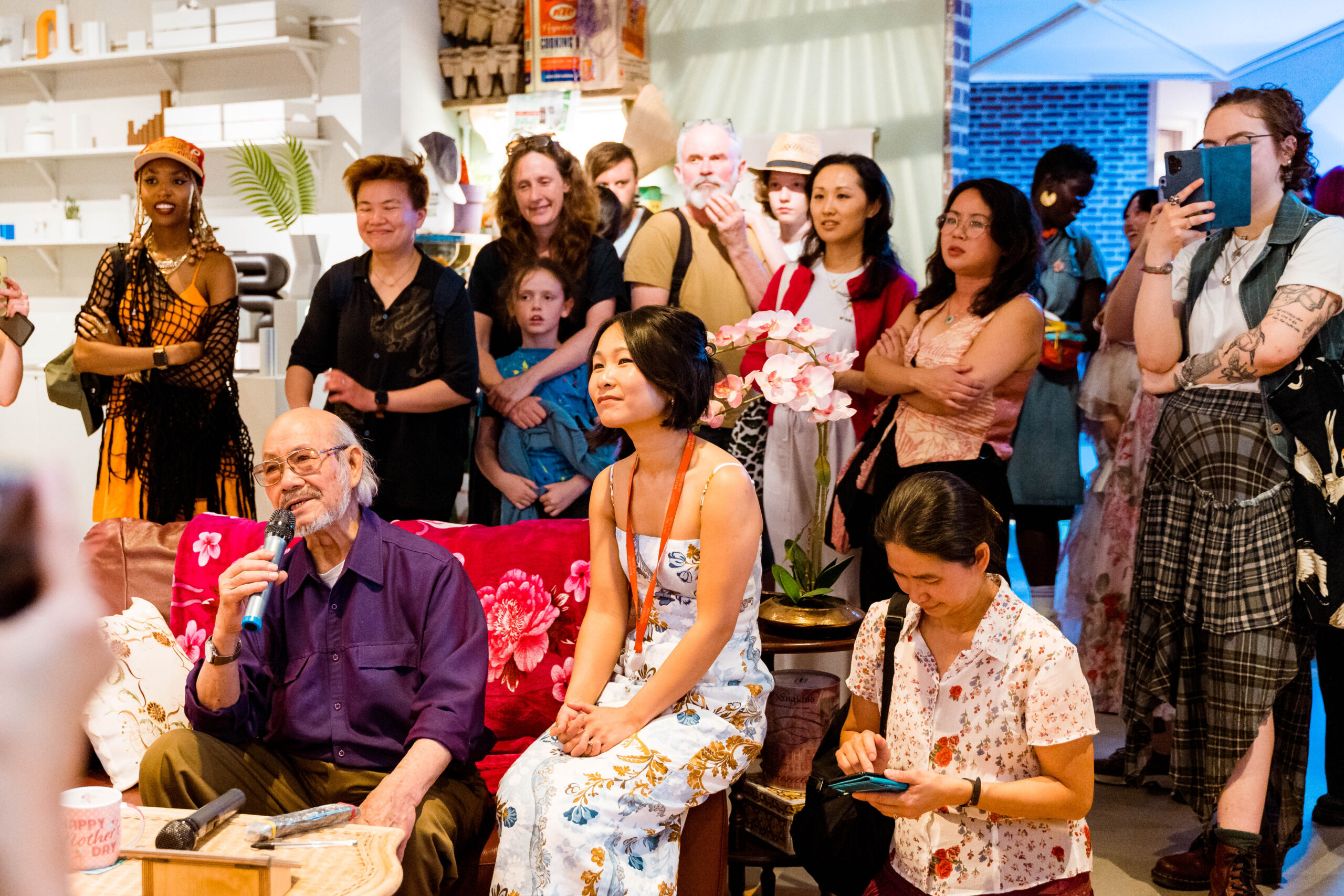 An older man sings karaoke on a sofa with a floral blanket over in A Terraced House in 2024 installation, while co-curator Anna Nguyen sits beside him and visitors listen and interact.