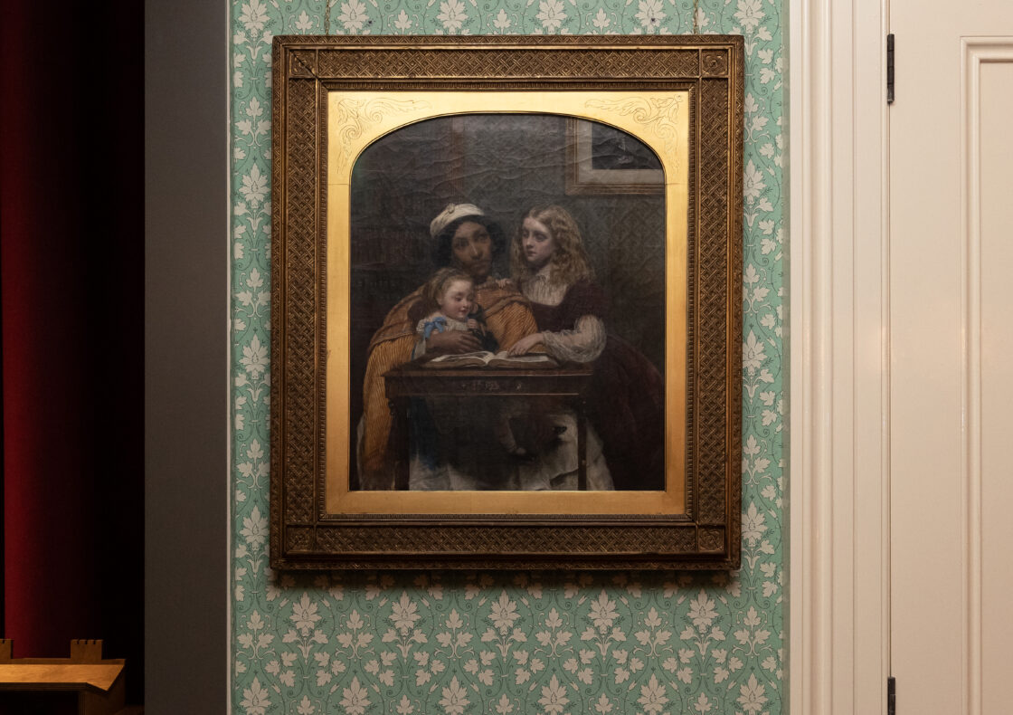 A framed painting hangs on floral-patterned wallpaper depicting a South Asian ayah—modelled by Jamaican-born Fanny Eaton—in an orange drape and white skirt, reading from a book to two white children, a toddler on her lap and an older girl leaning on the table.