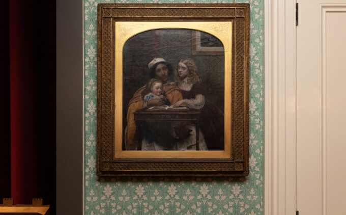 A framed painting hangs on floral-patterned wallpaper depicting a South Asian ayah—modelled by Jamaican-born Fanny Eaton—in an orange drape and white skirt, reading from a book to two white children, a toddler on her lap and an older girl leaning on the table.