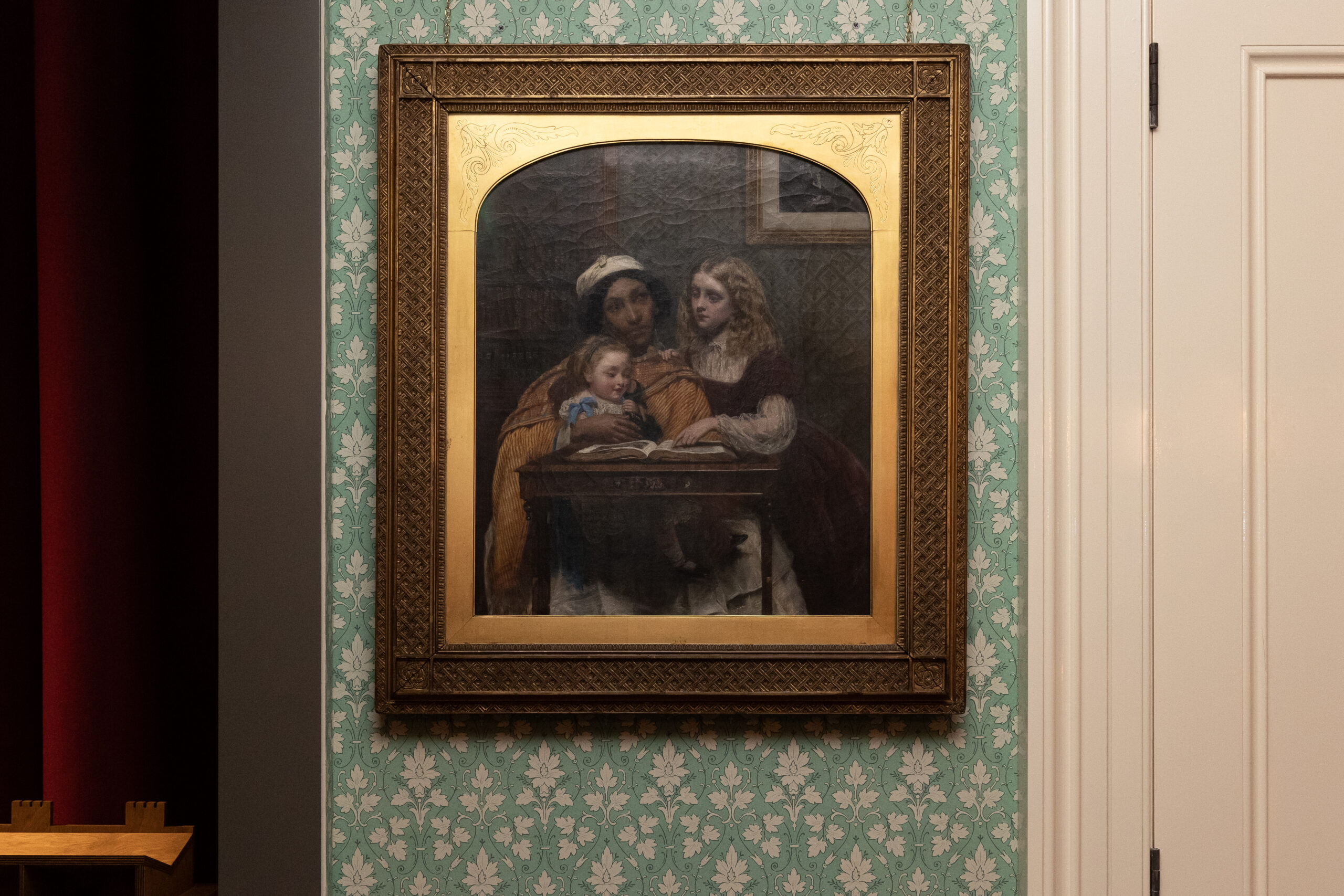 A framed painting hangs on floral-patterned wallpaper depicting a South Asian ayah—modelled by Jamaican-born Fanny Eaton—in an orange drape and white skirt, reading from a book to two white children, a toddler on her lap and an older girl leaning on the table.