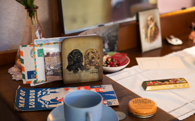 Various items rest on a vanity table in A Room Upstairs in 1956 at the Museum, including a Map of London, postcards and letters from Ireland, a blue teacup, and an ointment tin, among other objects, beneath a mirror.