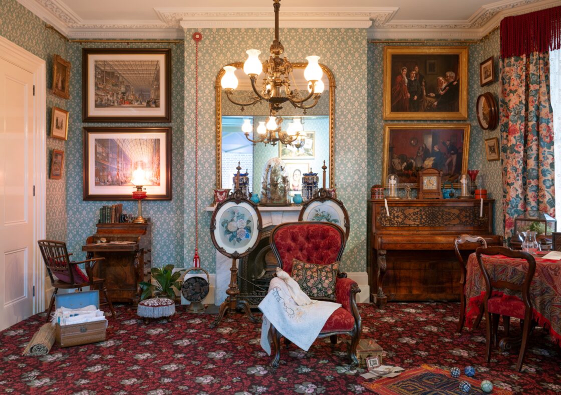 A richly furnished Victorian townhouse room (c.1878) with mint green patterned wallpaper and red carpet, featuring a red upholstered armchair draped with embroidery beside a fireplace topped by twin ceramic vases, framed artwork, a carved wooden piano, a tea table, and in the left bottom corner is an ayah's trunk of Indian textiles.
