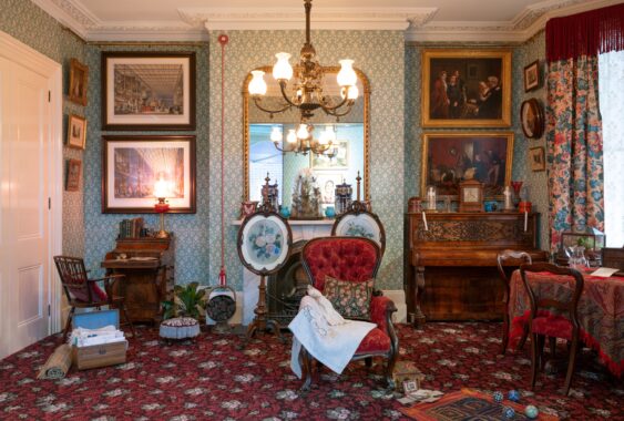 A richly furnished Victorian townhouse room (c.1878) with mint green patterned wallpaper and red carpet, featuring a red upholstered armchair draped with embroidery beside a fireplace topped by twin ceramic vases, framed artwork, a carved wooden piano, a tea table, and in the left bottom corner is an ayah's trunk of Indian textiles.