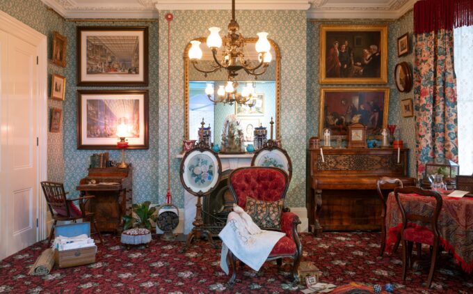 A richly furnished Victorian townhouse room (c.1878) with mint green patterned wallpaper and red carpet, featuring a red upholstered armchair draped with embroidery beside a fireplace topped by twin ceramic vases, framed artwork, a carved wooden piano, a tea table, and in the left bottom corner is an ayah's trunk of Indian textiles.