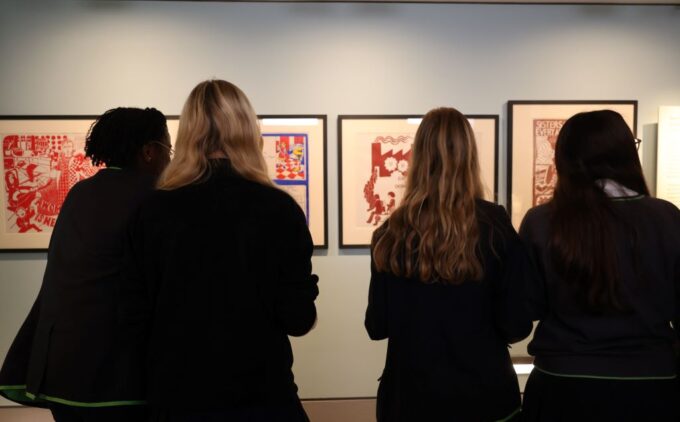 Four students in school uniforms stand shoulder to shoulder, their backs to the camera, viewing a row of predominantly red printmaking artworks framed on a pale blue gallery wall.