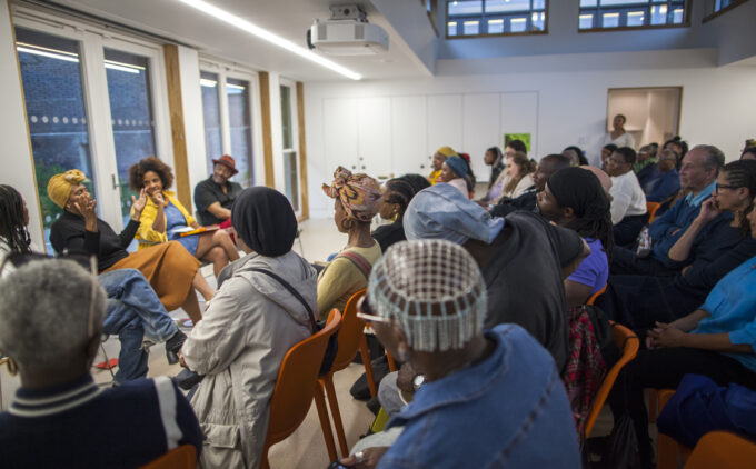 A packed audience sits in orange chairs watching a panel talk in a room with large windows.