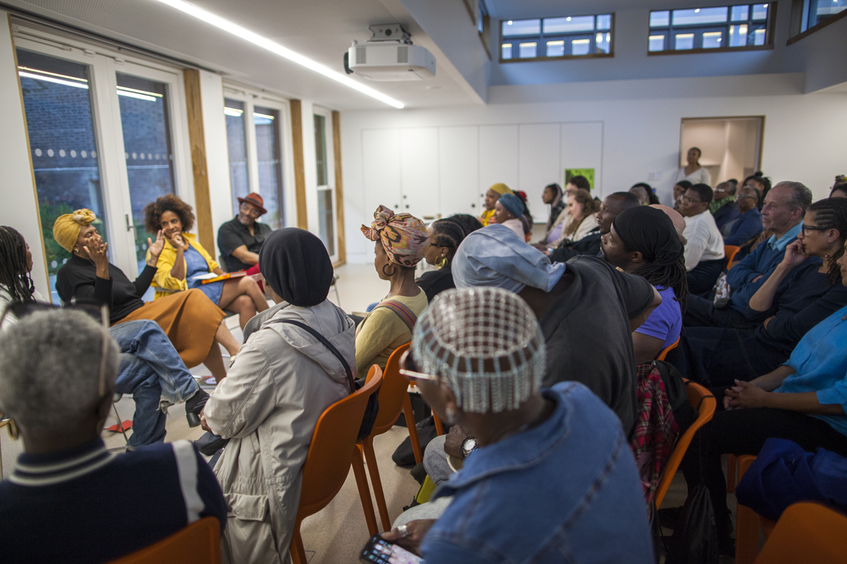 A packed audience sits in orange chairs watching a panel talk in a room with large windows.