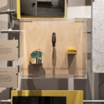 A close up shot of a clear acrylic display box mounted on a plywood panel, containing three objects: a colourful ceramic teapot in the left; a single metal screwdriver with translucent black handle in the centre; and a yellow rack holding multiple screw bits. Around this box are two caption panels describing the objects, and scaffolding poles and other exhibition panels and frame the installation.