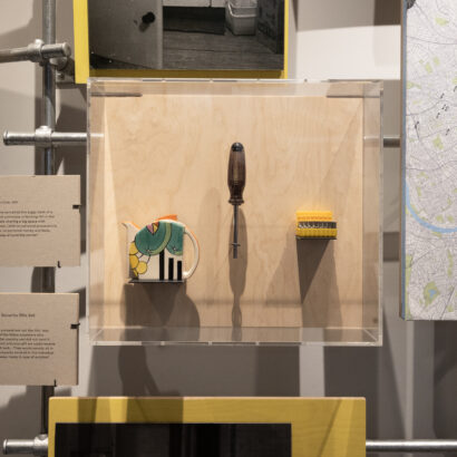 A close up shot of a clear acrylic display box mounted on a plywood panel, containing three objects: a colourful ceramic teapot in the left; a single metal screwdriver with translucent black handle in the centre; and a yellow rack holding multiple screw bits. Around this box are two caption panels describing the objects, and scaffolding poles and other exhibition panels and frame the installation.