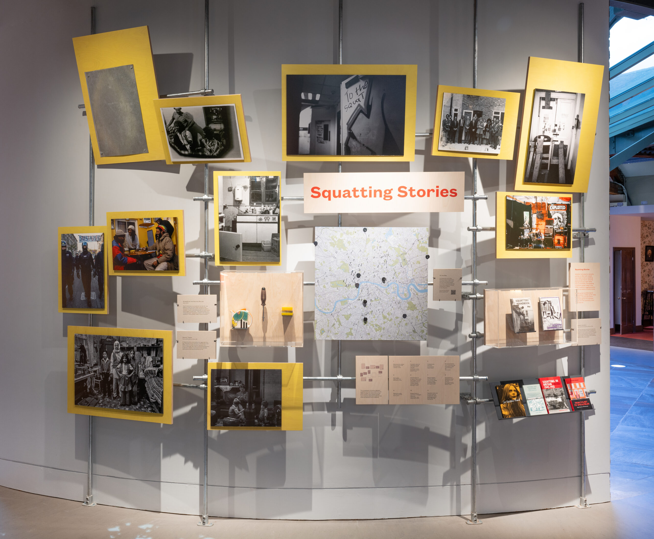 A display titled 'Squatting Stories' featuring multiple framed photographs in yellow frames arranged on a white wall with informational placards, a central map, and various historical images related to squatting and housing activism.