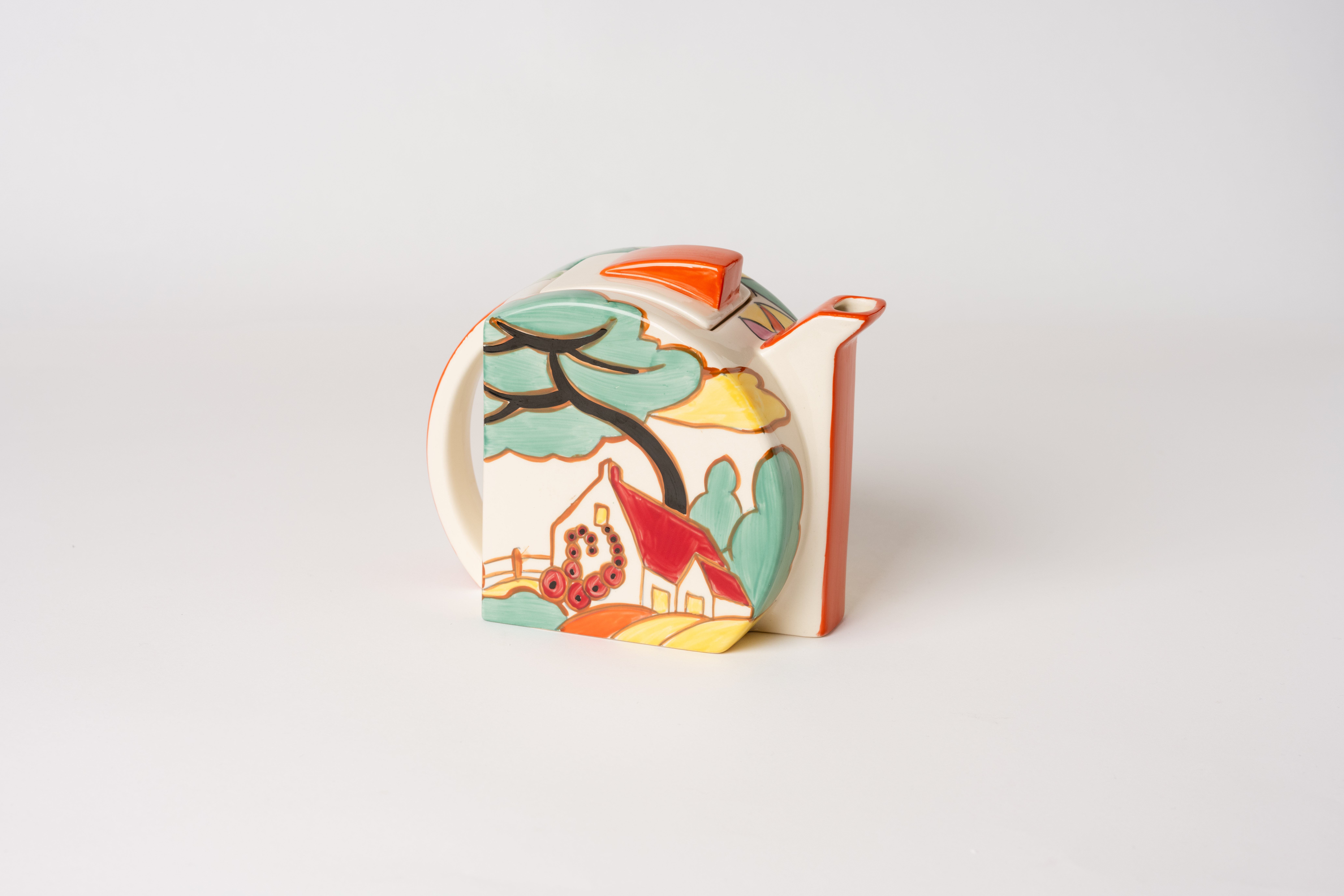 A ceramic teapot with a decorative design featuring a house and trees in green, yellow, and red.