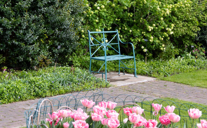 A blue, green bench sits among greenery with two brick paths leading up to it. In the forefront are pink flowers.