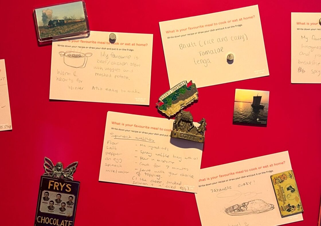 Multiple white recipe cards with handwritten recipes pinned to a red panel by souvenir magnets - a Fry’s Chocolate fairy, a camel and pyramids, Stirling Castle, a steam train, and a sunset sailboat - each answering “What is your favourite meal to cook or eat at home?”