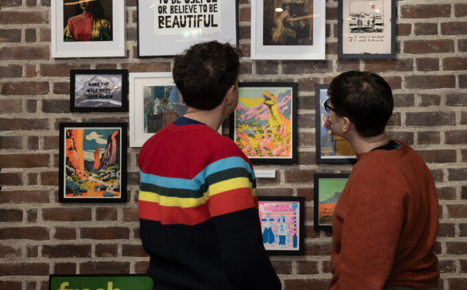 two people look at a wall of prints and framed art while talking