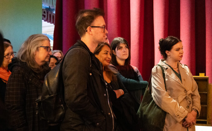 A diverse group of visitors stands before red curtains in a gallery, listening intently during a curator's guided tour.