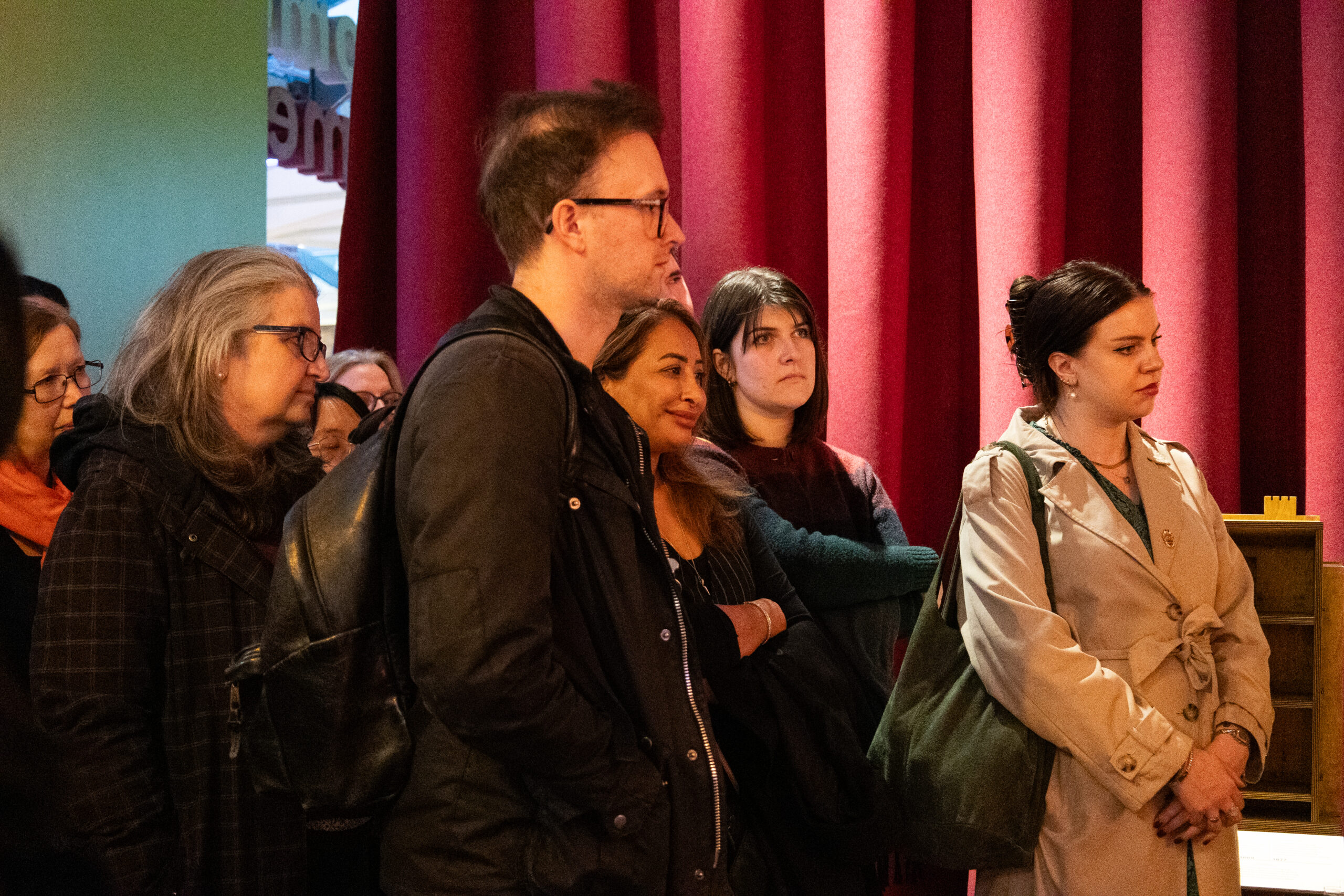 A diverse group of visitors stands before red curtains in a gallery, listening intently during a curator's guided tour.