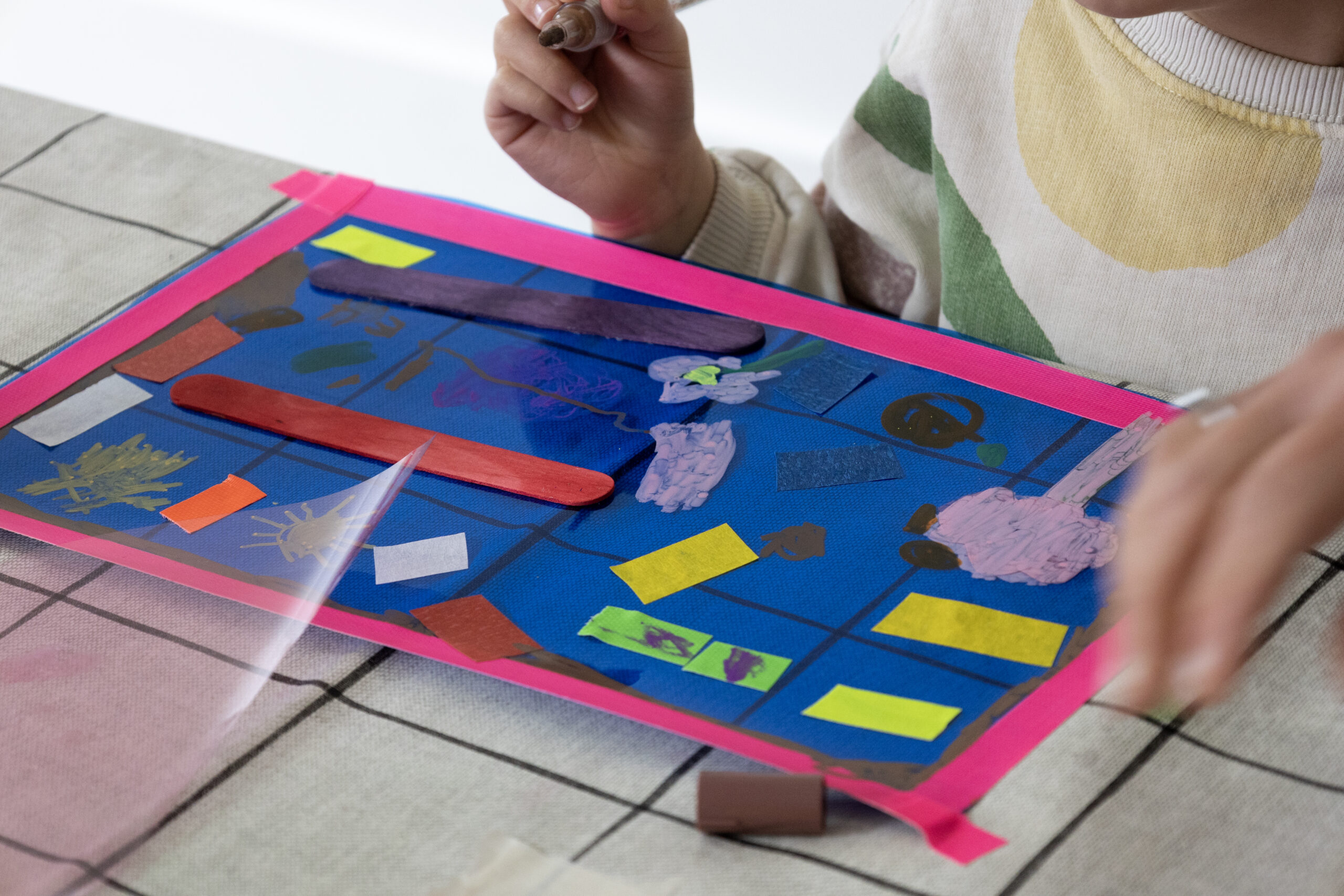 A child’s hand holds a brown marker above a translucent plastic overlay taped with pink masking tape to a table; on it are two painted popsicle sticks, small rectangles of coloured paper, and doodles in various hues.