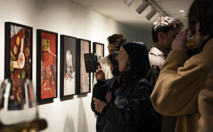 Visitors at the private view of “Still Living” lean in to examine a row of framed artworks under gallery track lighting, some capturing images on their phones and glasses raised in a toast.
