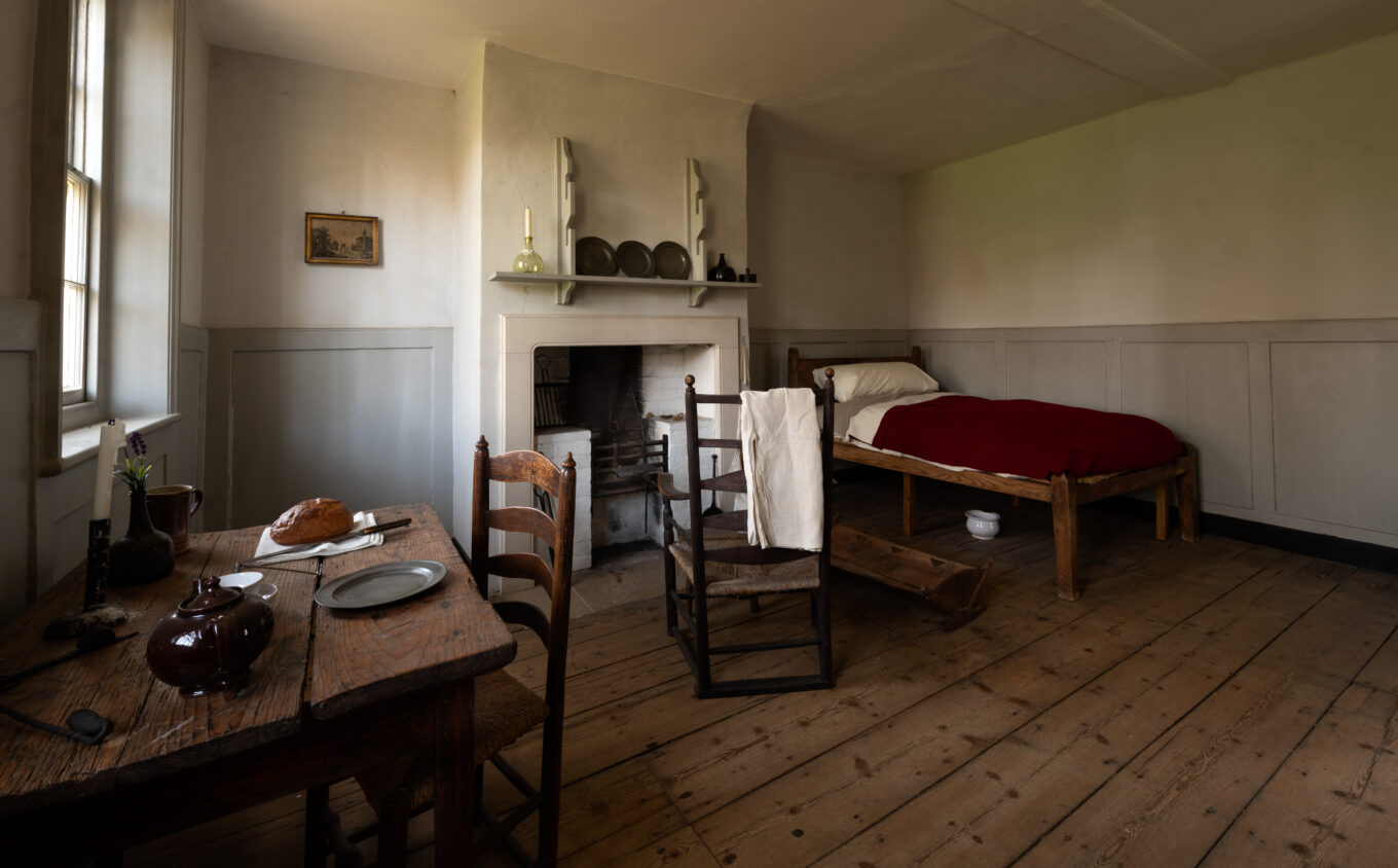 A simple almshouse room with a bed, fireplace and table with wooden floors