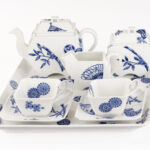 White porcelain tea set on a white background: a teapot, sugar bowl, and milk jug, plus two square cups with matching saucers, all decorated with indigo-blue patterns of bamboo, flowers and fans, arranged on a rectangular tray.