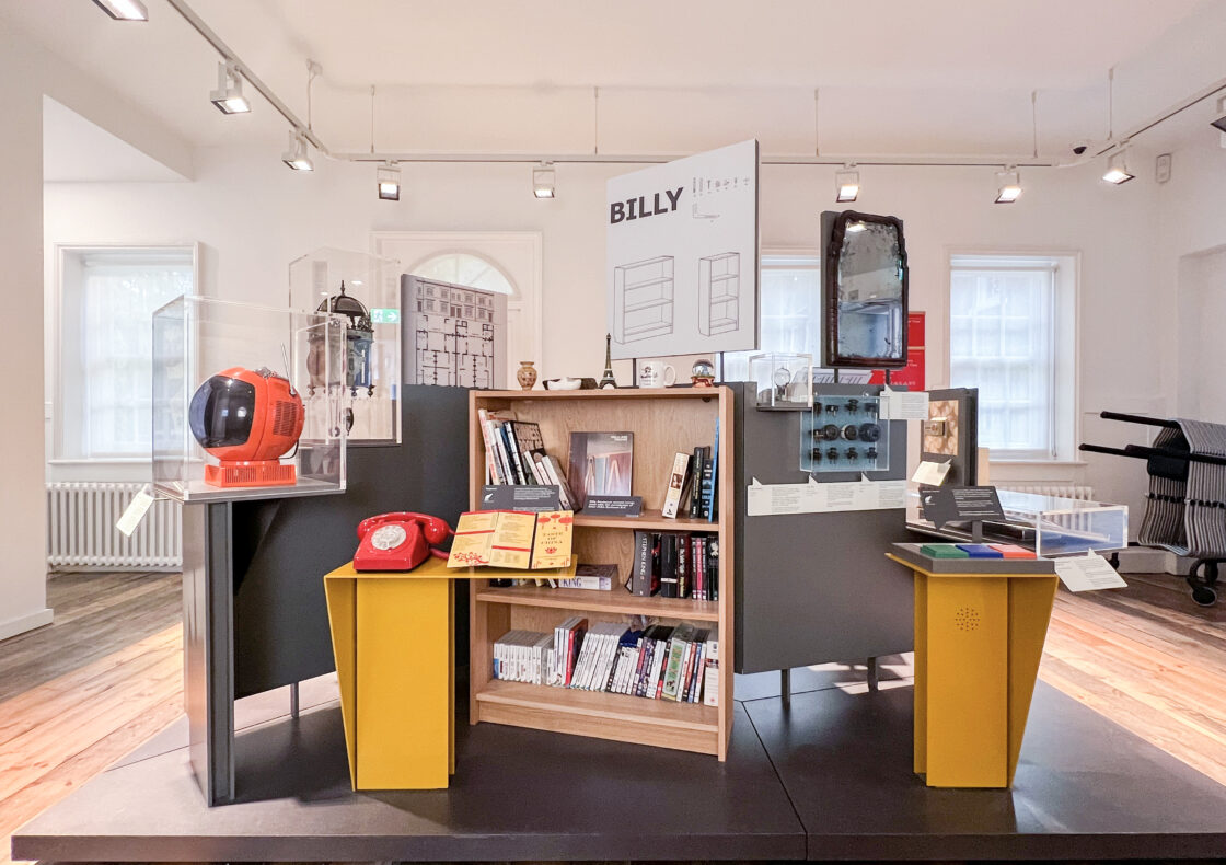 A standing display in a museum of objects including a red telephone, IKEA 'Billy' bookshelf, and mirror
