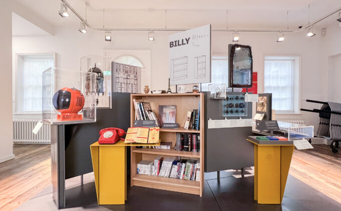 A standing display in a museum of objects including a red telephone, IKEA 'Billy' bookshelf, and mirror