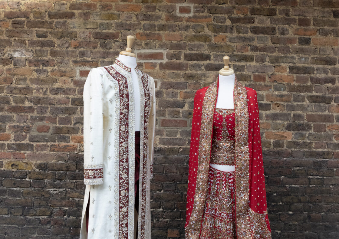Wedding attire displayed on mannequins against a brick wall - a cream-colored men's sherwani with burgundy embroidery and matching pants, alongside an ornate red and gold women's lehenga with intricate beadwork and embellishments.