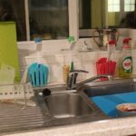 Neatly organised kitchen sink area: a stainless-steel double sink with an integrated draining board on the left, holding a dish rack with a green chopping board and white drying tray. Against the tiled windowsill stand three colour-coded brush holders (blue, green, red) each with a matching scrub brush, a bottle of Fairy lemon dish soap; inside the right basin sits a blue plastic tub with a white bowl that has some sauces still inside and purple-handled spoon.