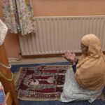 A woman kneels on a burgundy-and-cream prayer mat in a carpeted room, hands raised in supplication. She wears a mustard-coloured headscarf draped over a pale patterned fabric before a radiator beneath floral curtains. To the left, the wooden bedframe and the mattress are visible, with a white knitted beanie hanging from its post.