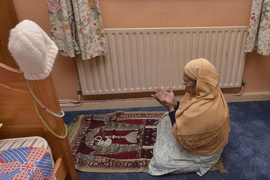A woman kneels on a burgundy-and-cream prayer mat in a carpeted room, hands raised in supplication. She wears a mustard-coloured headscarf draped over a pale patterned fabric before a radiator beneath floral curtains. To the left, the wooden bedframe and the mattress are visible, with a white knitted beanie hanging from its post.