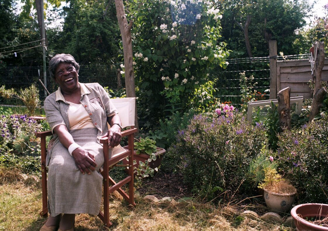 An elderly woman sits on the left in a wooden folding chair in a sunlit garden, laughing with her hands in her lap. She wears a pale striped skirt and matching jacket over a cream top, with open toe shoes. Behind her, potted plants and flowering shrubs, including tall bushes with white blossoms, stand before a wooden fence.
