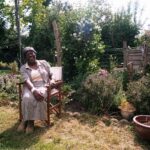 An elderly woman sits on the left in a wooden folding chair in a sunlit garden, laughing with her hands in her lap. She wears a pale striped skirt and matching jacket over a cream top, with open toe shoes. Behind her, potted plants and flowering shrubs, including tall bushes with white blossoms, stand before a wooden fence.