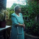 An elderly woman stands in a garden in the shade, facing right. On the left, there is a large potted plant resting on a table, and behind her are dense leafy shrubs that climb a wooden trellis.