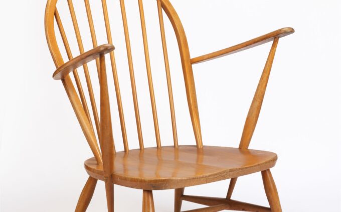A light-wood lounge chair with a wide gently scooped seat supported on four splayed legs and stretchers. Its backrest is formed by a tall curved hoop frame with evenly spaced vertical spindles, and its thin, angled arm rests extend outward from the top of two front spindles.