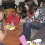A family plays a tabletop game on a brown ottoman in a living room. On the left, a man in a striped polo shirt leans forward; in the centre, a young girl in a red dress reaches for a game piece; on the right, a woman in a navy-and-white striped top and red scarf watches with hands poised. A green wicker chair, blue sofa, and stereo system fill the background.